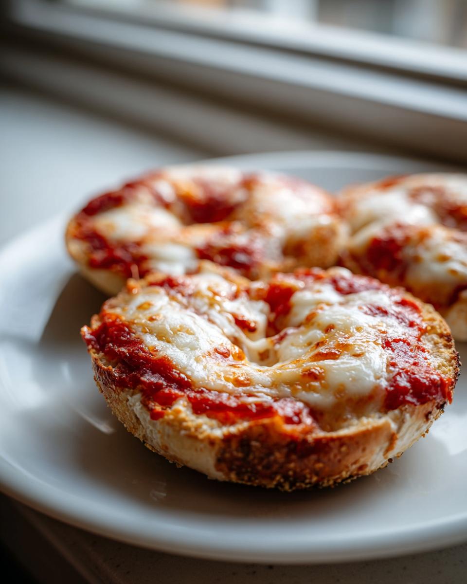 Close-up of three freshly made pizza bagels topped with melted mozzarella cheese and tomato sauce on a white plate.