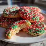 A plate of soft christmas sprinkle sugar cookies covered in red, green, and white nonpareils, one cookie is broken open.