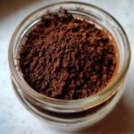 A close-up, top-down view of rich brown powder, representing homemade hot cocoa mix, filling a small glass jar.