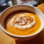 A close-up of vibrant orange butternut squash soup topped with a swirl of cream and a sprinkle of spice.