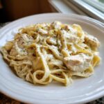 Close-up of creamy chicken alfredo pasta with fettuccine noodles and herbs on a white plate.