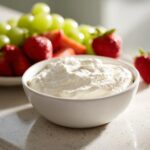 A bowl of creamy white fruit dip sits in the foreground, with fresh strawberries and green grapes blurred in the background.