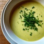 A close-up overhead shot of creamy, pale green leek soup garnished generously with fresh, chopped chives.