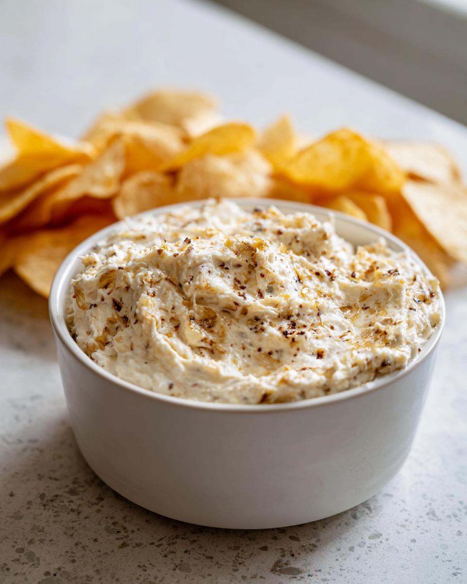 A white bowl filled with creamy onion dip topped with spices, with potato chips blurred in the background.