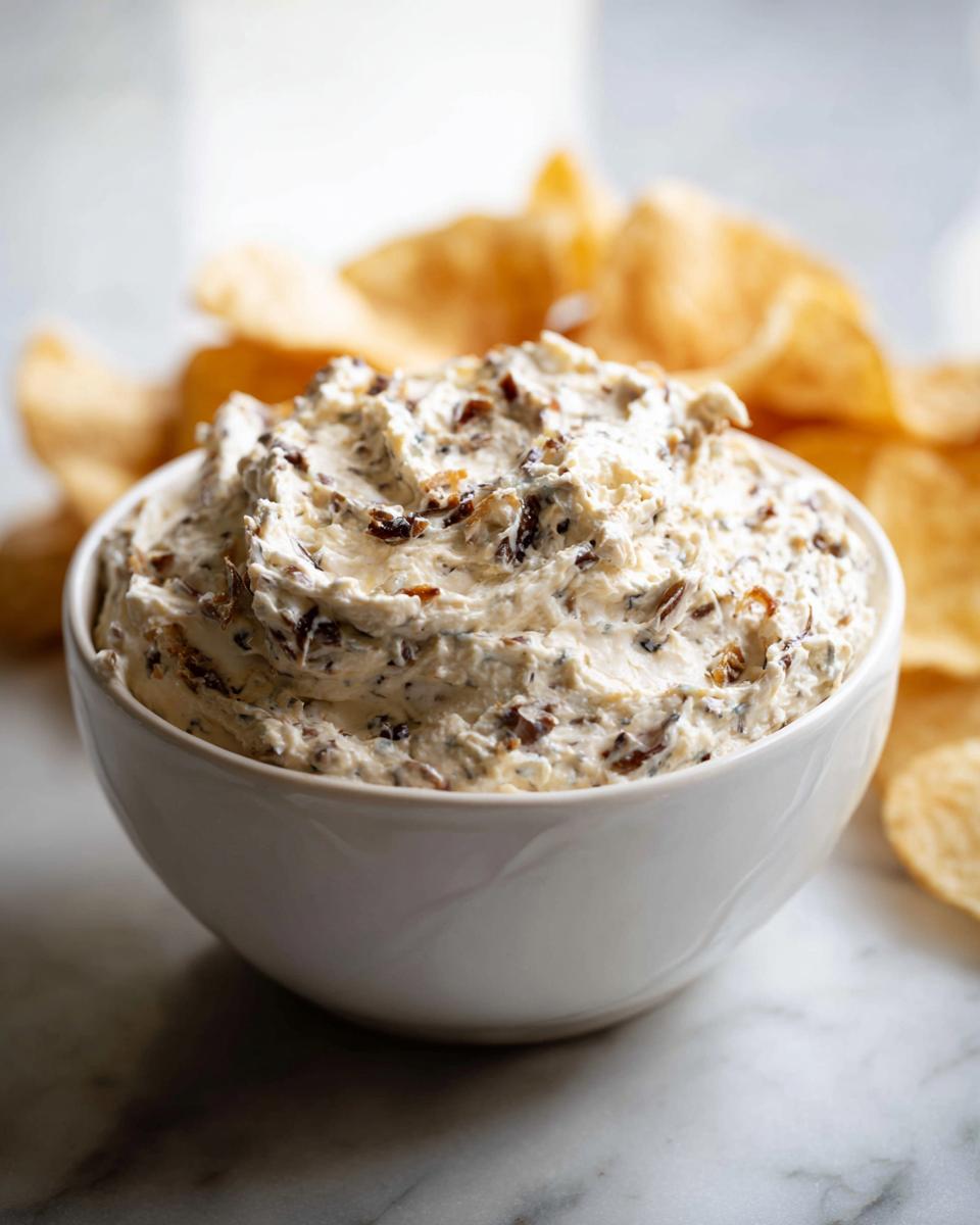 A close-up of a white bowl filled with creamy onion dip mixed with caramelized onions, served next to potato chips.