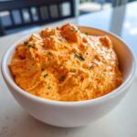 Close-up of a creamy, orange-hued boat dip sprinkled with red spices and green herbs in a white bowl.