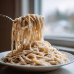 A fork lifts long strands of pasta coated thickly in creamy vegan alfredo sauce from a white bowl.