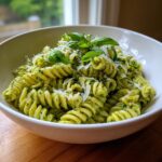 A white bowl filled with creamy pesto pasta (fusilli) topped with shaved Parmesan cheese and fresh basil leaves.