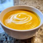 Close-up of a white bowl filled with bright orange pumpkin soup, garnished with a swirl of cream.