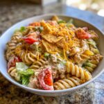 A close-up of a creamy taco pasta salad featuring rotini pasta, tomatoes, lettuce, cheese, and crushed tortilla chips.