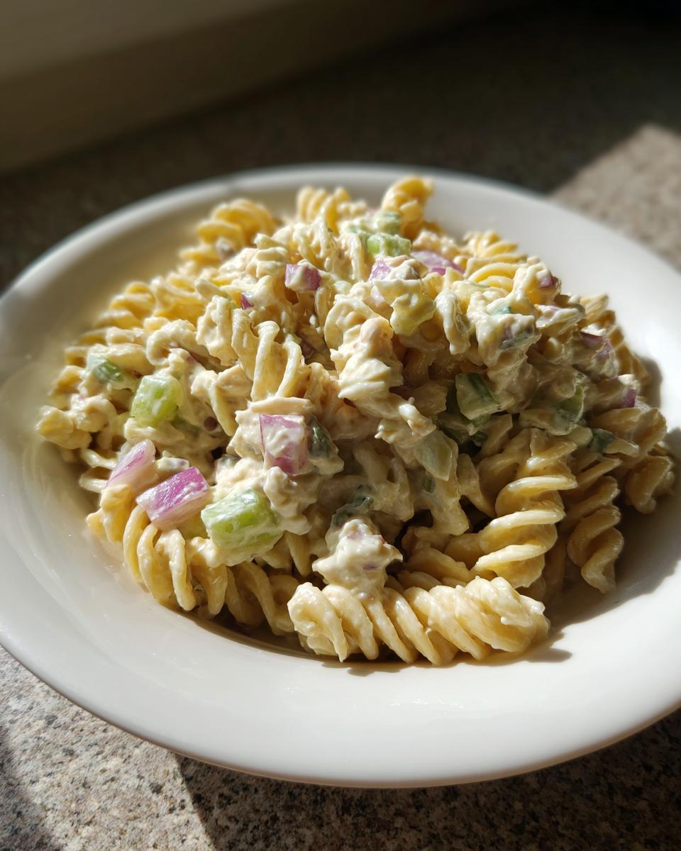 A close-up serving of creamy Tuna pasta salad made with rotini pasta, celery, and red onion in a white bowl.