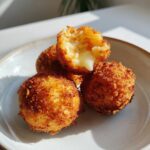 Close-up of crispy, golden brown rice balls, one broken open revealing a melted cheese center.