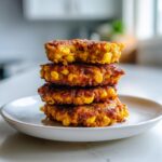 A tall stack of four golden-brown, crispy corn fritters piled on a white plate.