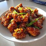 Close-up of crispy, glazed orange chicken recipe pieces topped with sesame seeds and sliced green onions.