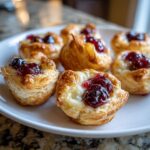 Close-up of several warm, flaky brie bites topped with melted brie and dark cherry jam.