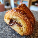Close-up of a flaky, golden brown chocolate croissant cut open to show melted chocolate filling.