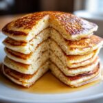Close-up of a tall stack of fluffy discard pancakes with a slice cut out, drizzled with syrup.