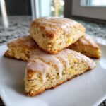 Close-up of four fluffy lemon scones stacked on a white plate, drizzled with a thick lemon glaze and sprinkled with zest.