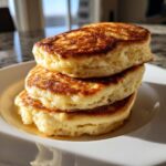 A close-up stack of three thick, golden-brown sourdough pancakes drizzled with syrup on a white plate.
