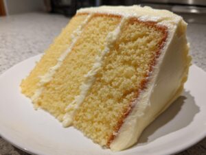 Close-up of a moist, fluffy slice of vanilla cake recipe with thick white buttercream frosting layers.