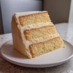Close-up of a moist, three-layer slice from a vanilla cake recipe, covered in white frosting.