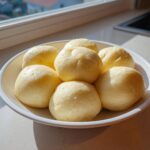 A white bowl filled with several smooth, pale yellow balls of freshly made fufu, sitting on a countertop near a window.