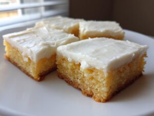 Four squares of thick, moist sugar cookie bars topped with creamy white frosting on a white plate.