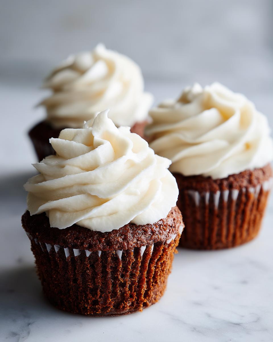 Close-up of three rich, dark gingerbread cupcakes topped with tall swirls of white cream cheese frosting.