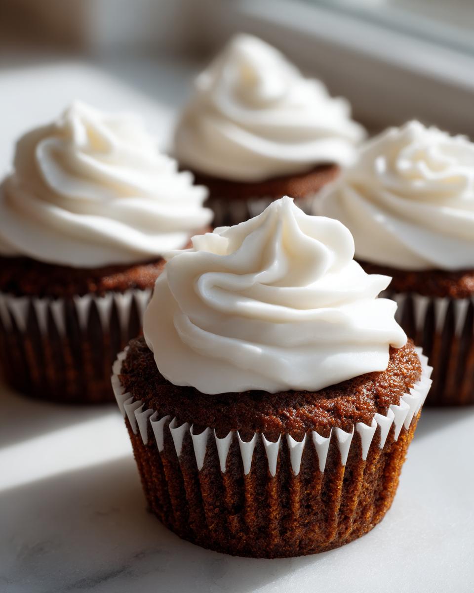 A close-up of one perfectly frosted gingerbread cupcake with white cream cheese frosting, with three others blurred in the background.