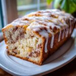 A close-up of a freshly baked apple fritter bread loaf drizzled generously with white icing, resting on a white plate.