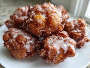 A close-up of several golden-brown, glazed apple fritter mounds piled on a white plate.