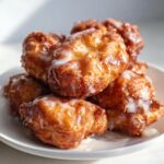 A close-up stack of golden brown, freshly fried apple fritter treats drizzled with sweet white glaze.