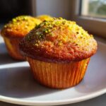 Close-up of a golden-brown pistachio muffin topped with coarse sugar and chopped green pistachios.