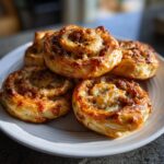 A close-up of four golden brown, cheesy sausage pinwheels stacked on a white plate.