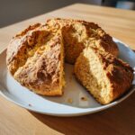 A freshly baked, round loaf of golden brown soda bread, sliced on a light plate.