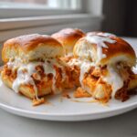 Close-up of gooey buffalo chicken sliders overflowing with shredded chicken and white sauce on soft rolls.