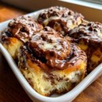 Four freshly baked chocolate cinnamon rolls covered in thick, glossy chocolate frosting, sitting in a white baking dish.
