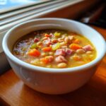 A close-up of a white bowl filled with homemade bean soup featuring white beans, diced ham, carrots, and celery.
