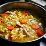Close-up view of rich broth chicken noodle soup with egg noodles, shredded chicken, and sliced carrots simmering in a pot.