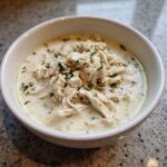 A close-up of a white bowl filled with creamy homemade cream of chicken soup, topped generously with shredded chicken and fresh herbs.