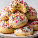 A stack of soft Italian ricotta cookies topped with white glaze and colorful rainbow sprinkles, one cookie has a bite taken out.