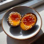 Two halves of a marinated egg with a jammy, deep orange yolk, sprinkled with white sesame seeds, served in a small bowl.