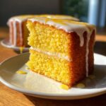 Close-up of a moist slice of bright yellow lemon cake recipe topped with white icing and lemon zest.