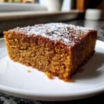 Close-up of a moist slice of dark brown applesauce cake dusted with powdered sugar on a white plate.