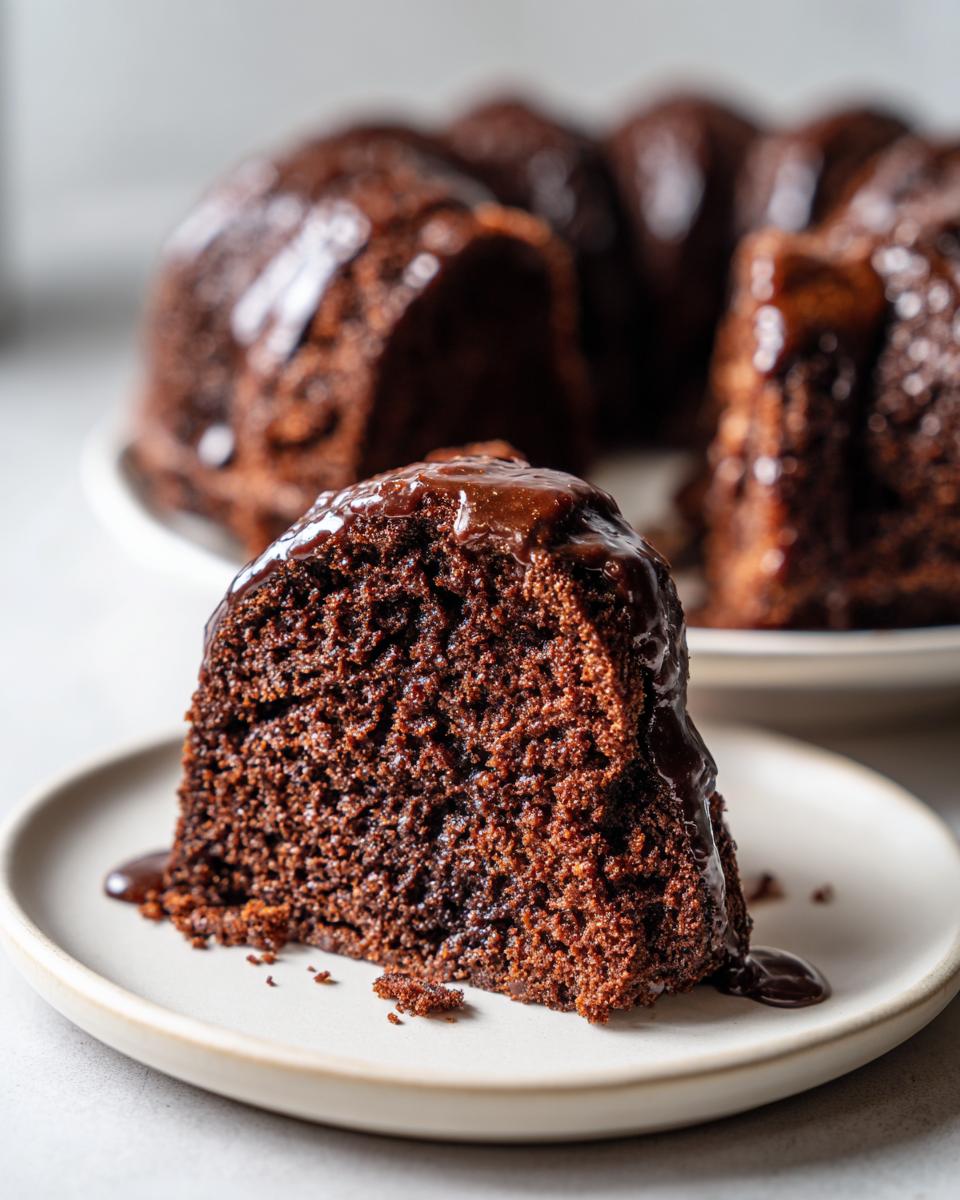 Close-up of a moist slice of dark gingerbread cake topped with rich chocolate glaze, with the full bundt cake blurred in the background.