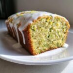 A close-up of a loaf of lemon zucchini bread, topped with a thick white glaze and lemon zest.