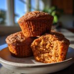 A stack of three moist sweet potato muffins, one broken open to show the texture, sitting on a light plate.