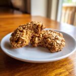Two soft, oatmeal-textured applesauce cookies on a white plate, one broken in half.