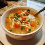 A close-up of a warm bowl of penicillin soup featuring shredded chicken, diced carrots, Israeli couscous, and fresh parsley.