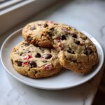 Three freshly baked peppermint chocolate chip cookies stacked slightly on a white plate near a window.